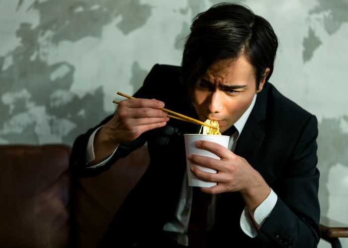 A man in a suit eating noodles. He looks slightly nervous.